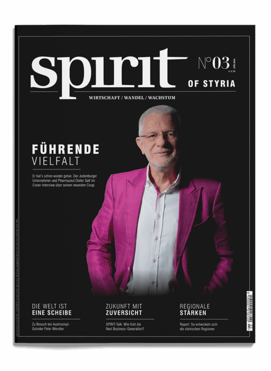 Ein lächelnder älterer Mann mit Brille und weißem Bart trägt eine leuchtend rosa Anzugsjacke über einem weißen Hemd. Er steht selbstbewusst mit einer Hand in der Tasche vor einem schwarzen Hintergrund auf dem Cover des Magazins "spirit of Styria".