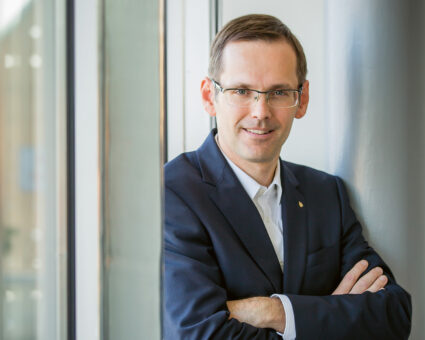 Ein Mann mit kurzen braunen Haaren und Brille, einem dunklen Anzug und weißem Hemd, steht mit verschränkten Armen und lächelt in der Nähe eines Fensters mit Tageslicht.