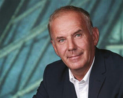 Ein Mann mittleren Alters mit kurzen, hellen Haaren trägt eine dunkle Anzugsjacke und ein weißes Hemd und schaut leicht lächelnd in die Kamera. Im Hintergrund ist ein abstraktes blaues und grünes Muster zu sehen.