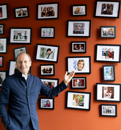 Ein lächelnder Mann in einer dunklen Jacke steht vor einer orangefarbenen Wand, die mit gerahmten Fotos von verschiedenen Personen und Ereignissen bedeckt ist, und zeigt mit seiner rechten Hand auf eines der Bilder.
