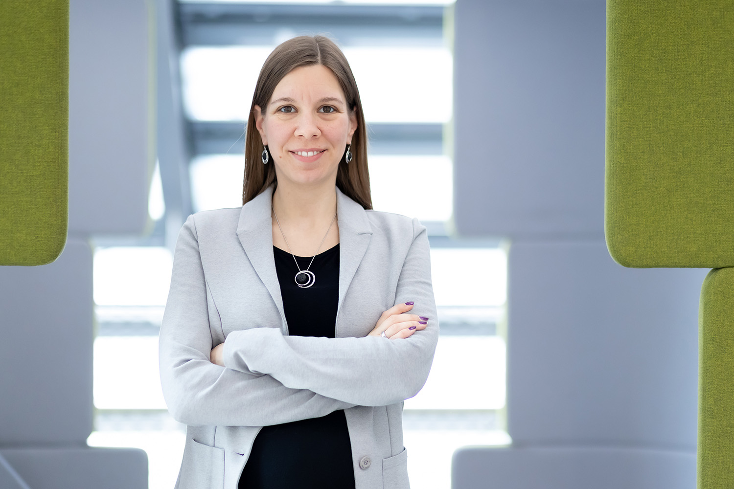 Eine Frau mit langen braunen Haaren, die einen hellgrauen Blazer und ein schwarzes Oberteil trägt, steht selbstbewusst mit verschränkten Armen in einem modernen, hell erleuchteten Büroraum mit grünen und grauen Tafeln.