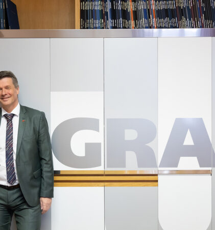 Ein Mann in grünem Anzug und gestreifter Krawatte steht lächelnd vor einer Wand, an der in großen Buchstaben "GRAZ" geschrieben steht und darüber ein Regal mit Büchern.