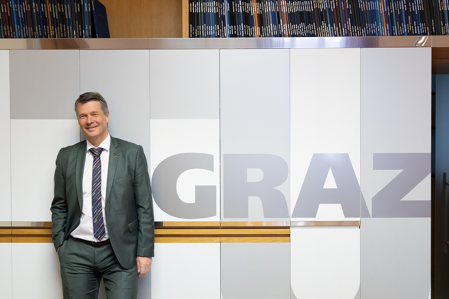 Ein Mann in grünem Anzug und gestreifter Krawatte steht lächelnd vor einer Wand, an der in großen Buchstaben "GRAZ" geschrieben steht und darüber ein Regal mit Büchern.