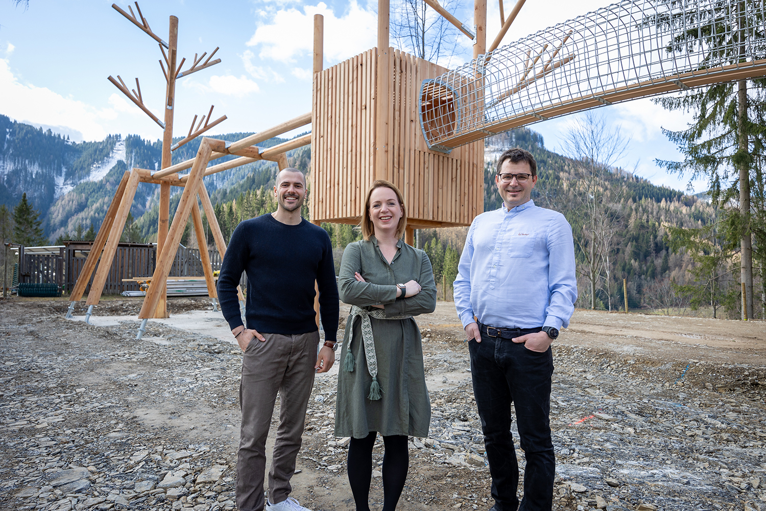 Drei Personen stehen lächelnd im Freien vor einem modernen Holzspielplatz mit einer Röhrenrutsche, umgeben von Bäumen und Bergen im Hintergrund.