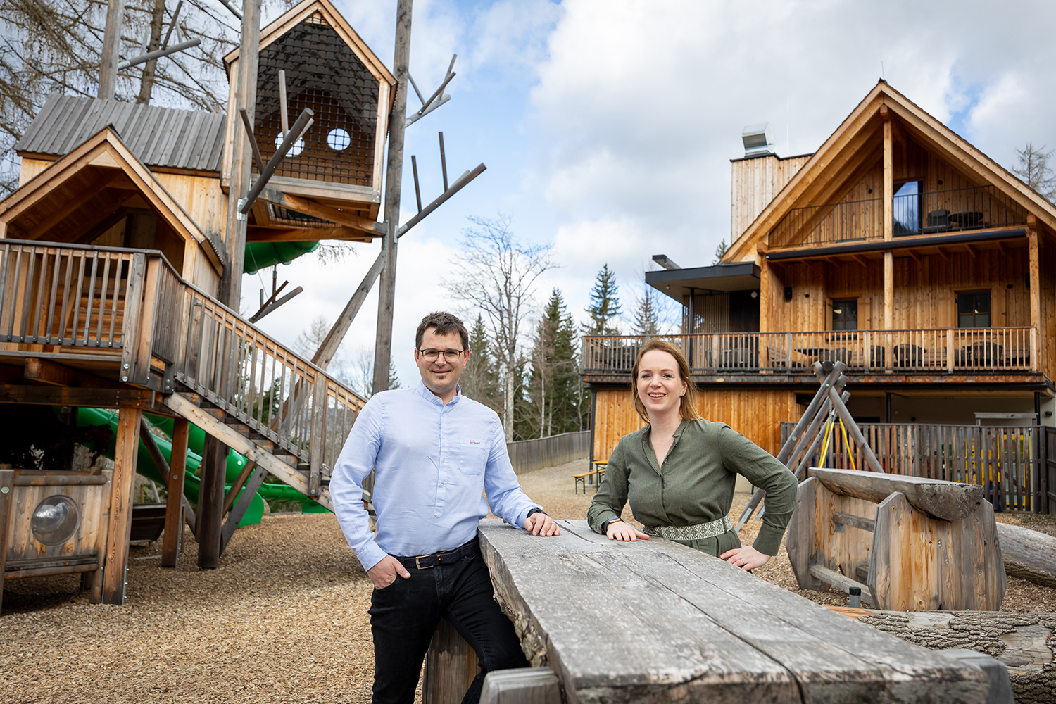 Zwei Personen, ein Mann und eine Frau, stehen lächelnd an einem rustikalen Holztisch in einem Außenbereich mit großen hölzernen Baumhäusern und Hütten im Hintergrund, umgeben von Bäumen und Spielgeräten.