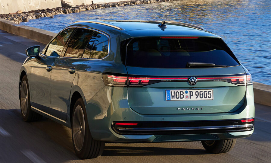 Ein grüner Volkswagen Passat Kombi fährt eine Uferstraße entlang, wobei sich das Sonnenlicht auf dem Wasser spiegelt und im Hintergrund Gebäude zu sehen sind.