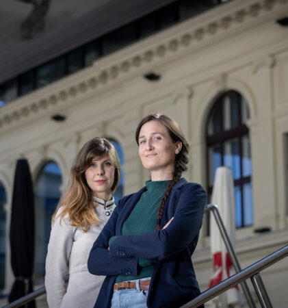 Zwei Frauen stehen selbstbewusst auf einer Freitreppe vor einem cremefarbenen Gebäude mit gewölbten Fenstern, die eine in einem hellen Mantel, die andere in einer dunklen Jacke, beide schauen in die Kamera.