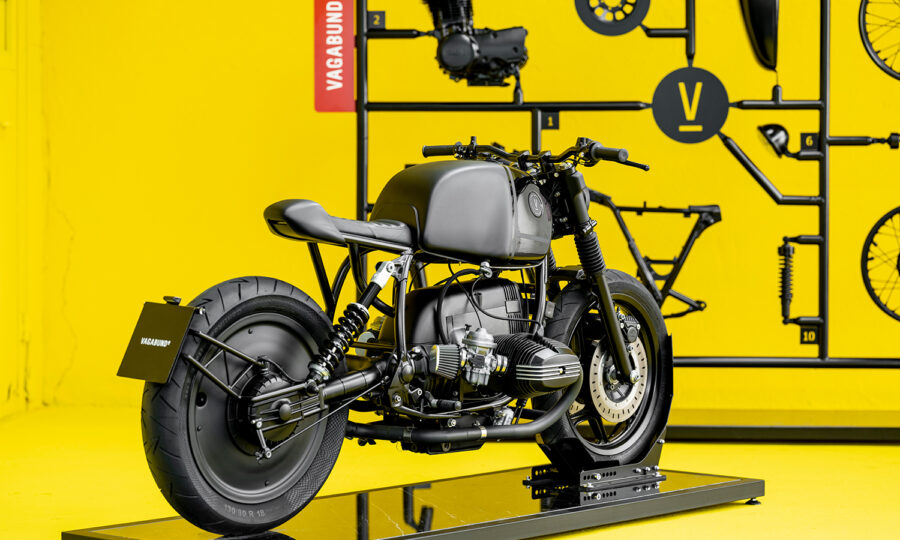 Ein schnittiges, mattschwarzes Custom-Motorrad steht auf einer reflektierenden Plattform vor einer leuchtend gelben Wand mit montierten Motorradteilen und einem roten Schild mit der Aufschrift "Vagabund".