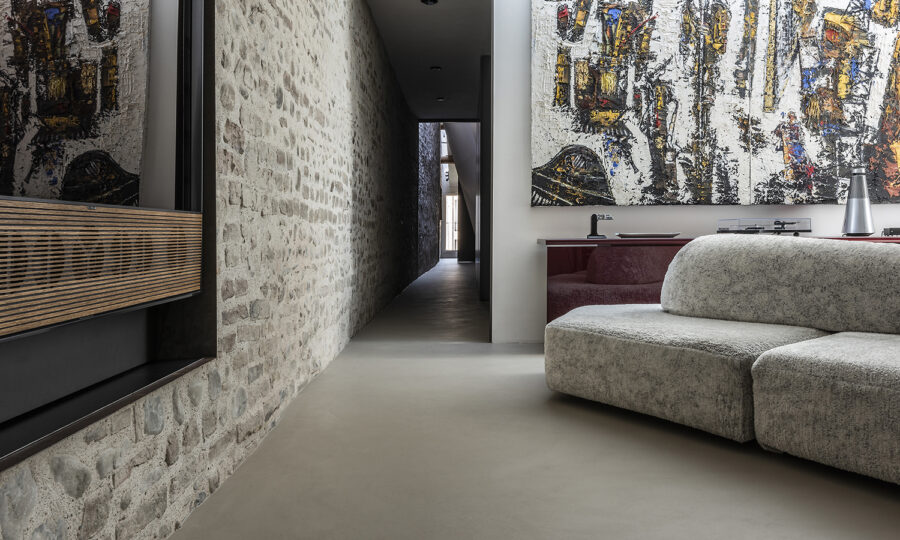 Ein moderner Wohnraum mit Steinwänden, einem strukturierten grauen Sofa, abstrakten Kunstwerken und einem langen Flur, der zu Tageslicht führt. Die Einrichtung ist minimalistisch, mit neutralen Tönen und zeitgenössischen Designelementen.