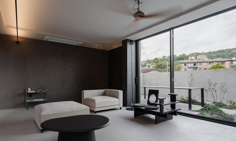 Minimalistisches Wohnzimmer in neutralen Tönen mit einem modernen Sessel, einer Ottomane, einem runden schwarzen Couchtisch und einem eleganten schwarzen Beistelltisch. Große, bodentiefe Fenster bieten einen Blick auf die Terrasse und das Grün draußen.