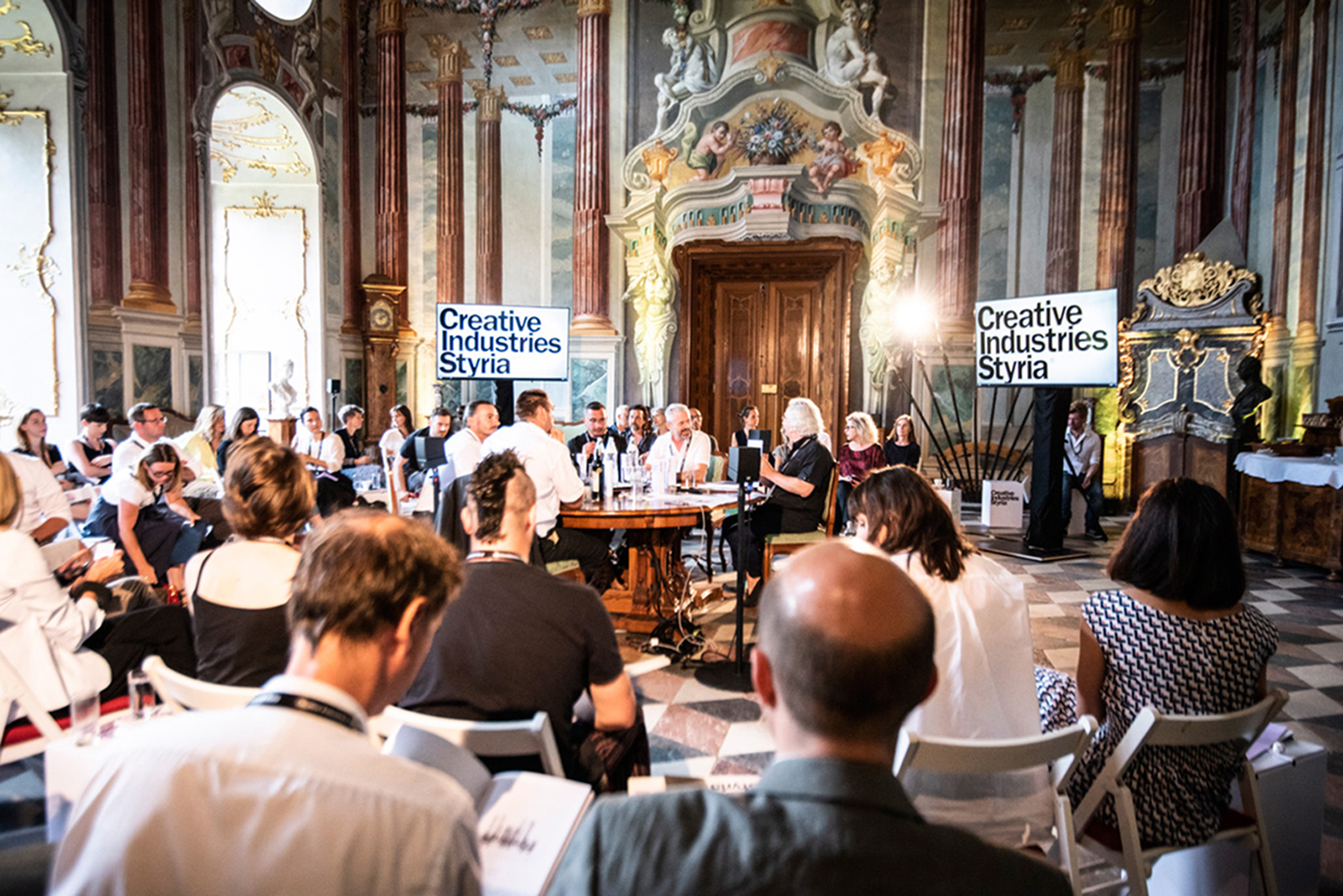 Eine Gruppe von Personen nimmt an einer Podiumsdiskussion in einem prunkvollen, barocken Saal mit großen Fenstern und dekorativen Wänden teil. Zwei Bildschirme zeigen "Creative Industries Styria". Das Publikum steht den Rednern an einem zentralen runden Tisch gegenüber.