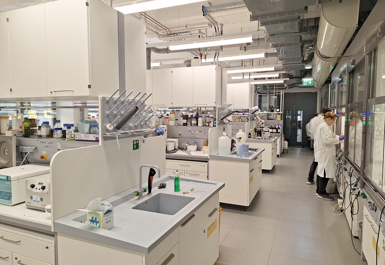 Ein modernes Labor mit weißen Arbeitsplätzen, Waschbecken und wissenschaftlichen Geräten. Zwei Personen in Laborkitteln arbeiten an einer Theke auf der rechten Seite, während auf anderen Bänken Flaschen, Werkzeuge und Zubehör stehen. Helle Beleuchtung und saubere Oberflächen überall.