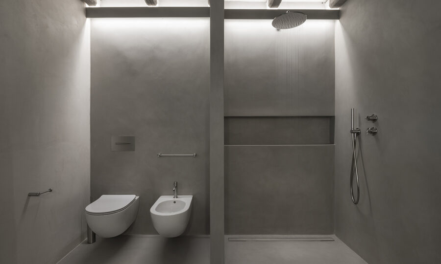 Minimalistisches, modernes Badezimmer mit grauen Wänden, einer wandmontierten Toilette und einem Bidet auf der linken Seite und einer offenen, begehbaren Dusche mit Regendusche und eingebautem Regal auf der rechten Seite, die durch eine Teilwand getrennt ist.