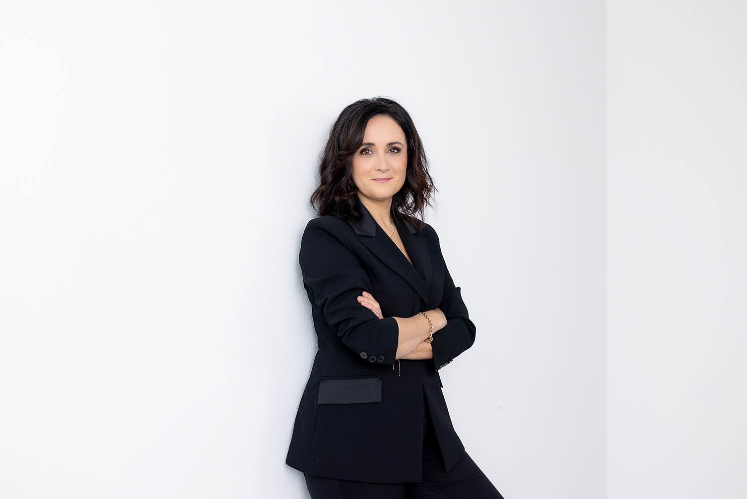 Eine Frau mit dunklem Haar und schwarzem Anzug steht mit verschränkten Armen vor einer weißen Wand und lächelt selbstbewusst in die Kamera.