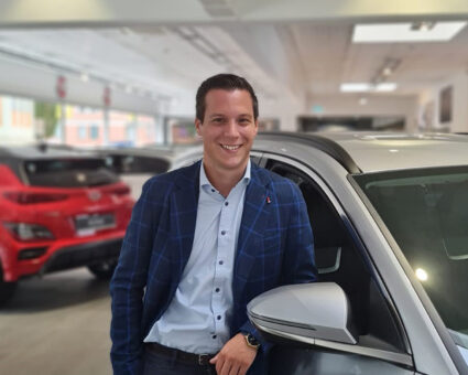 Ein Mann in einem blau karierten Blazer und weißem Hemd lächelt, während er sich an den Seitenspiegel eines silbernen Autos in einem hellen Autohaus mit anderen Fahrzeugen im Hintergrund lehnt.