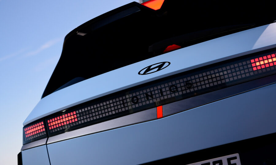 Nahaufnahme des Hecks eines silbernen Hyundai Ioniq 5-Elektroautos mit dem Hyundai-Logo, Rückleuchten im Pixel-Stil und einer Teilansicht des Nummernschilds vor blauem Himmel.