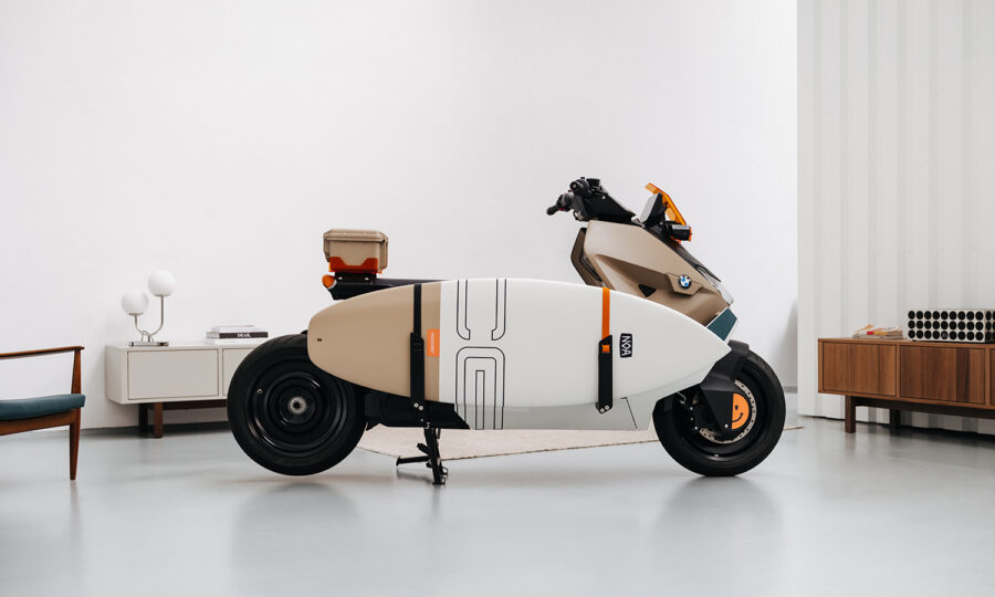 Ein moderner Elektroroller, an dessen Seite ein Surfbrett befestigt ist, steht in einem Innenraum auf einem hellen Boden neben minimalistischen Möbeln und weißen Wänden.