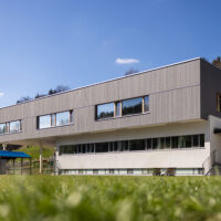Ein modernes, zweistöckiges Gebäude mit großen Fenstern und Holzvertäfelung im Obergeschoss, eingebettet in eine Wiese mit Hügeln und Bäumen im Hintergrund unter einem klaren blauen Himmel.