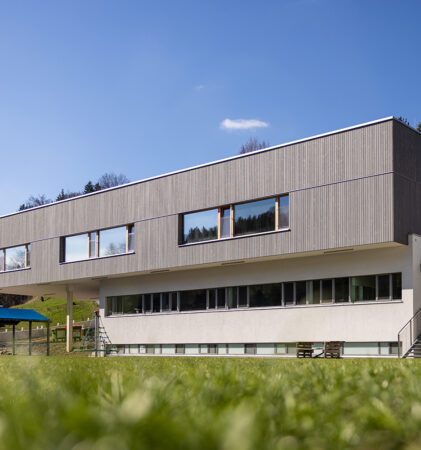 Ein modernes, zweistöckiges Gebäude mit großen Fenstern und Holzvertäfelung im Obergeschoss, eingebettet in eine Wiese mit Hügeln und Bäumen im Hintergrund unter einem klaren blauen Himmel.