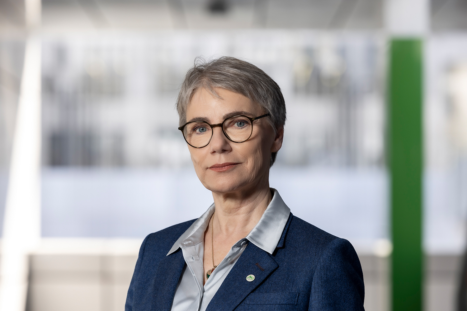 Eine selbstbewusste ältere Frau mit kurzen grauen Haaren und Brille trägt einen blauen Blazer über einer hellen Bluse und steht in einer modernen, hell erleuchteten Büroumgebung.