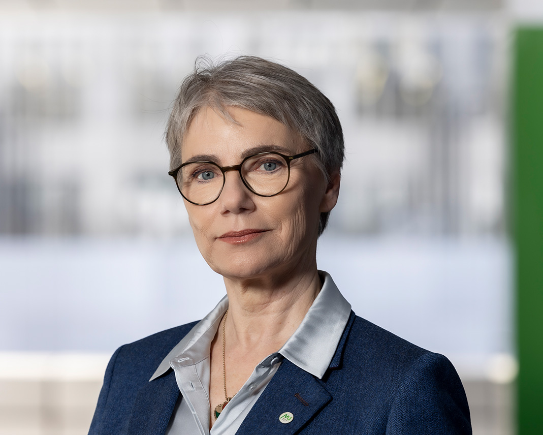 Eine Frau mittleren Alters mit kurzen grauen Haaren und Brille, die einen blauen Blazer und ein hellblaues Hemd trägt, steht vor einem verschwommenen, modernen Hintergrund. Sie blickt selbstbewusst in die Kamera.