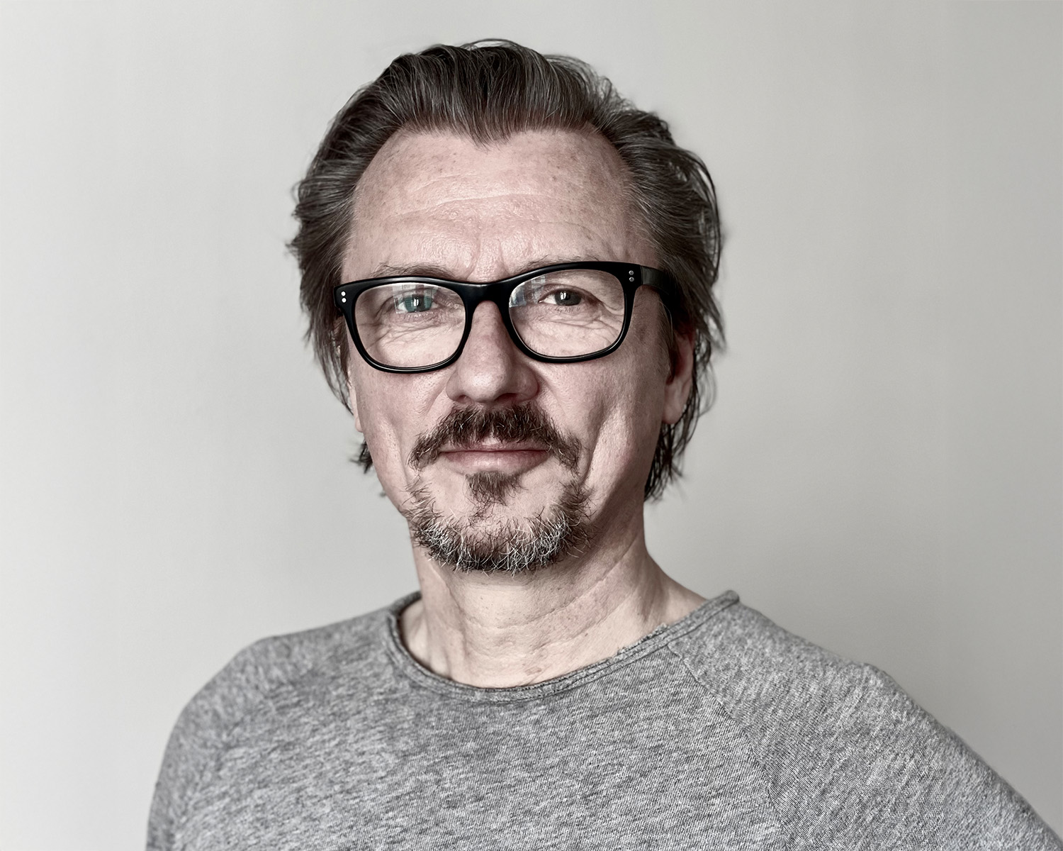 Ein Mann mittleren Alters mit heller Haut, kurzen braunen Haaren, Schnurrbart und Spitzbart, schwarzer Brille und grauem Hemd steht vor einem schlichten hellen Hintergrund und schaut mit neutralem Blick in die Kamera.