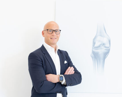 Ein glatzköpfiger Mann mit Brille und Anzug steht mit verschränkten Armen lächelnd vor einer weißen Wand mit einer anatomischen Abbildung eines Kniegelenks.