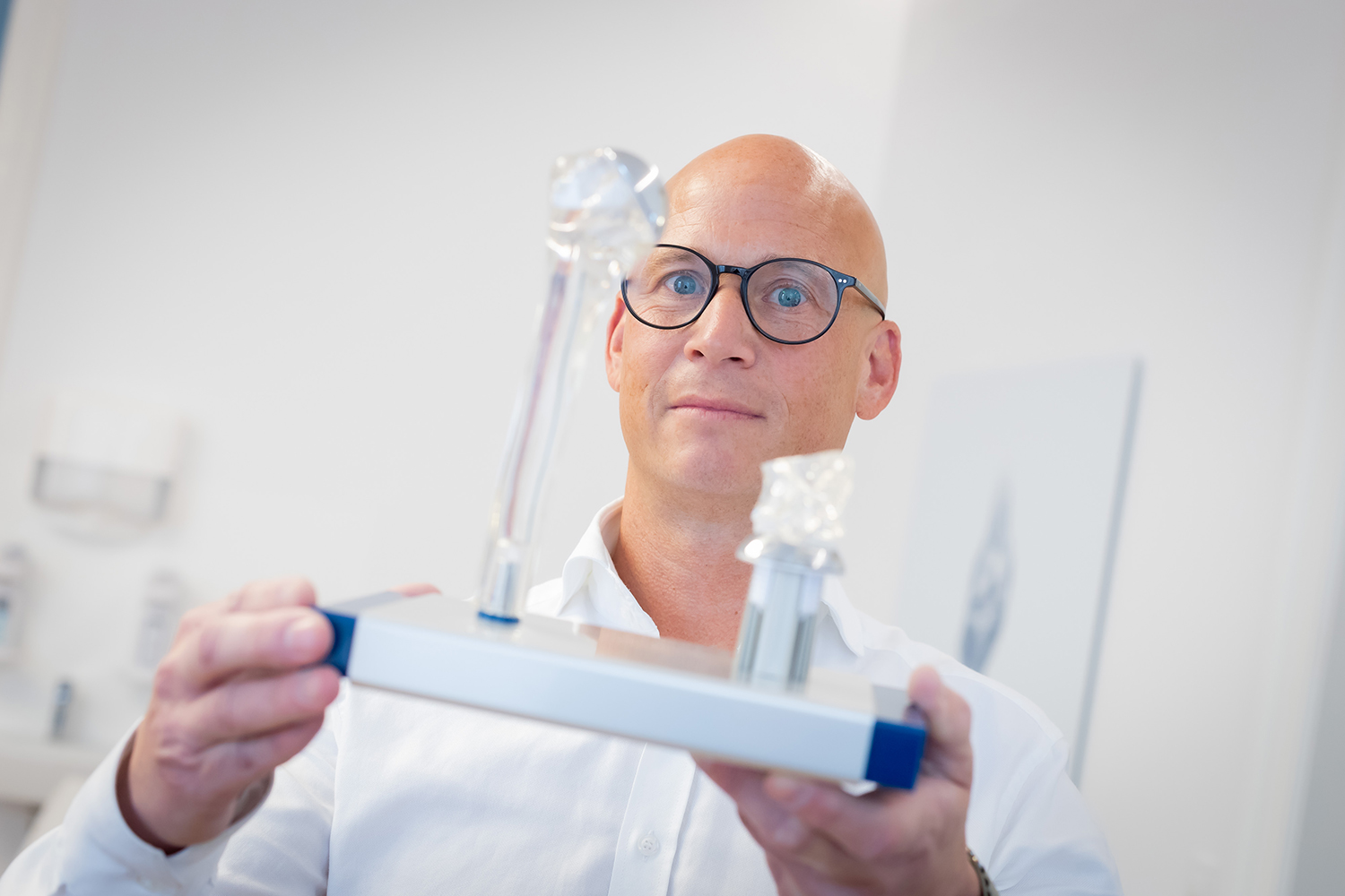 Ein glatzköpfiger Mann mit Brille und weißem Hemd hält ein Modell eines menschlichen Hüftgelenks hoch, wobei er die künstlichen Implantate in einer hellen medizinischen oder Laborumgebung in den Mittelpunkt stellt.