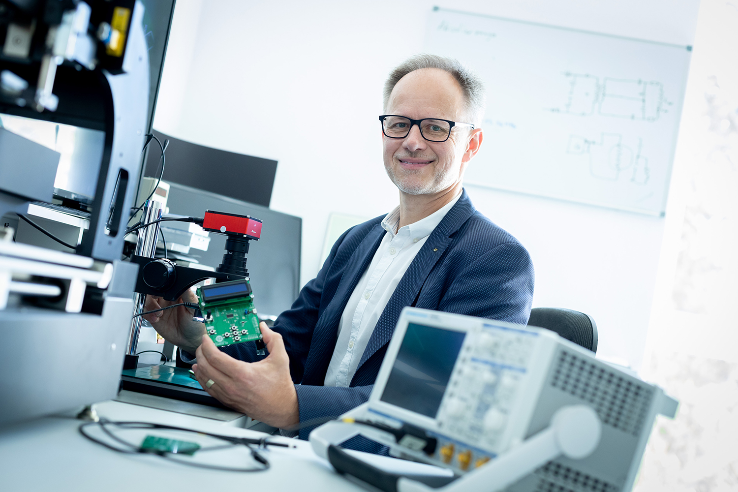 Ein Mann mit Brille und Anzugjacke sitzt an einem Schreibtisch mit elektronischen Geräten und hält eine grüne Platine in der Hand. Im Hintergrund sind Laborgeräte und ein Whiteboard mit Diagrammen zu sehen.