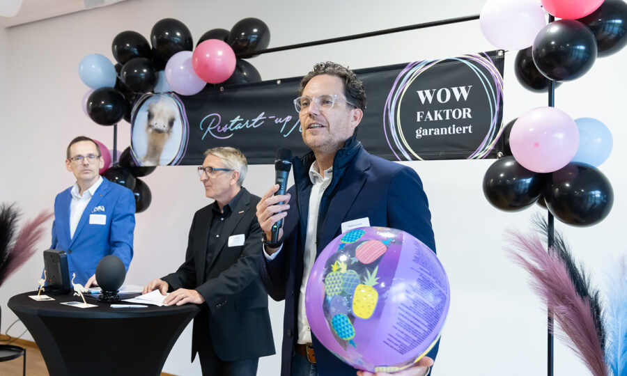 Drei Männer stehen vor einem Luftballon und einem Transparent mit der Aufschrift "WOW FAKTOR garantiert". Ein Mann hält einen bunten Strandball und spricht in ein Mikrofon, während die anderen an einem schwarzen Tisch mit Laptops und Mikrofonen stehen.