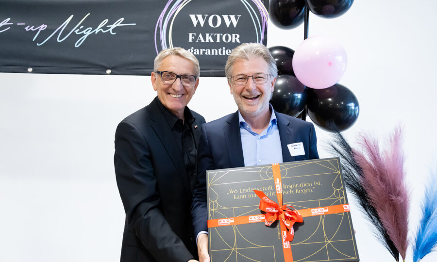 Zwei lächelnde Männer in Anzügen stehen bei einer Veranstaltung zusammen, einer von ihnen hält eine große, als Geschenk verpackte Schachtel mit einer roten Schleife. Hinter ihnen ist ein schwarzes Banner mit der Aufschrift "WOW FAKTOR" und Dekorationen mit schwarzen und rosafarbenen Luftballons zu sehen.