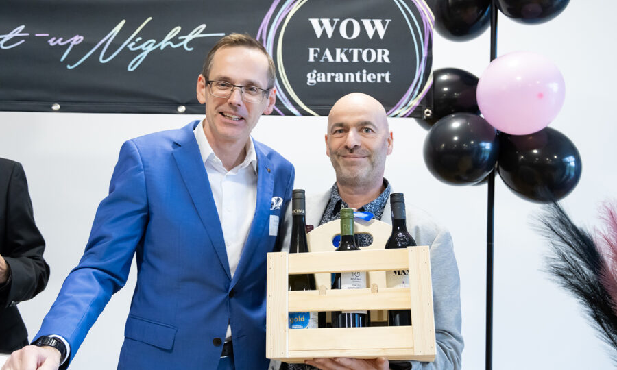 Zwei Männer in Anzügen lächeln bei einer Veranstaltung. Ein Mann hält eine Holzkiste mit vier Flaschen in der Hand. Hinter ihnen sind schwarze und rosa Luftballons und ein Schild mit der Aufschrift "WOW FAKTOR garantiert.