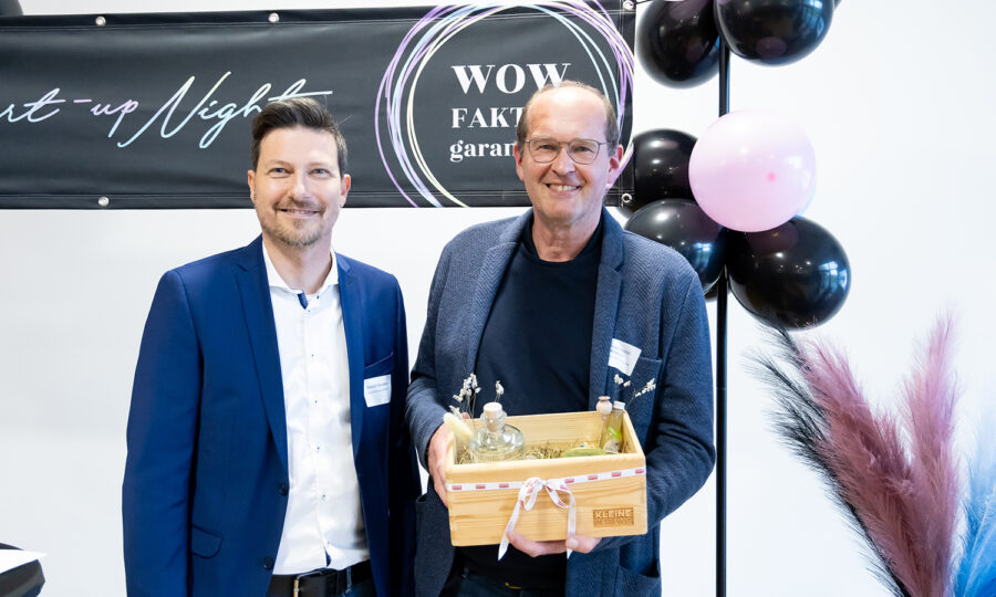 Zwei Männer in Geschäftskleidung stehen lächelnd vor einem "WOW FAKTOR garantiert"-Schild und schwarzen Luftballons; ein Mann hält eine hölzerne Geschenkbox mit Blumen und kleinen Gegenständen.