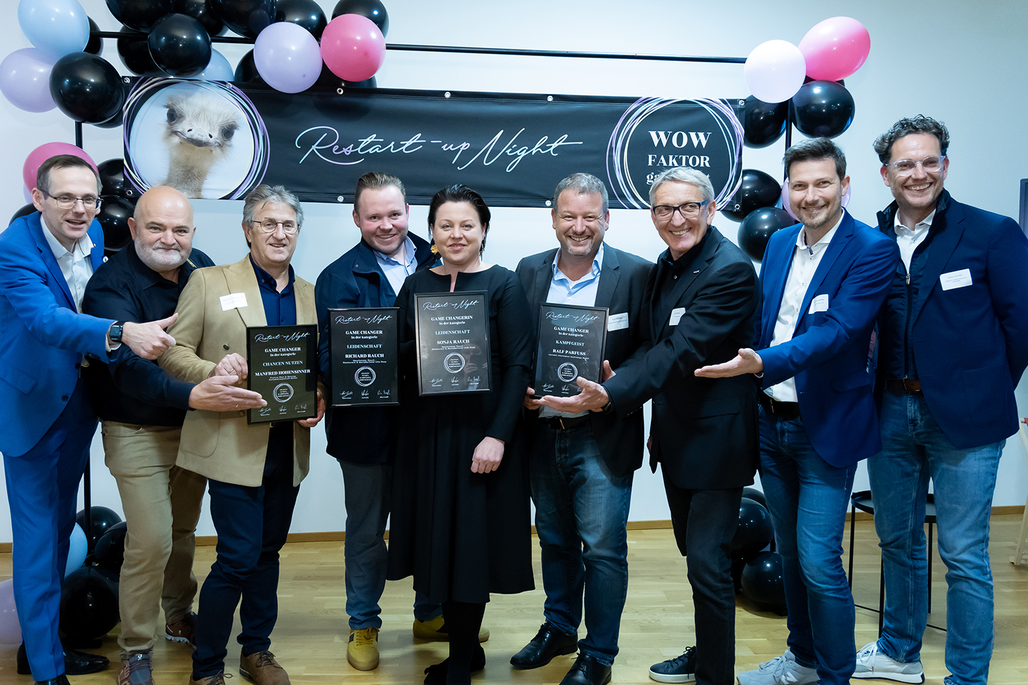 Eine Gruppe von acht Personen lächelt und posiert in einem Innenraum mit Preisplaketten in der Hand. Sie stehen vor einem Banner mit der Aufschrift "Restart-up Night" und "WOW FAKTOR", während schwarze, rosa und weiße Luftballons den Hintergrund zieren.