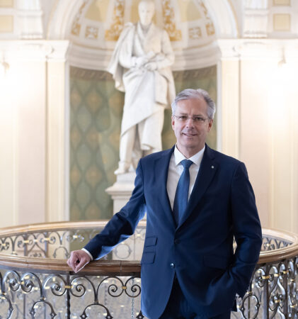 Ein Mann im Anzug steht an einem kunstvollen Geländer in einem eleganten Innenraum, mit einer großen weißen Statue und gewölbten architektonischen Details im Hintergrund.