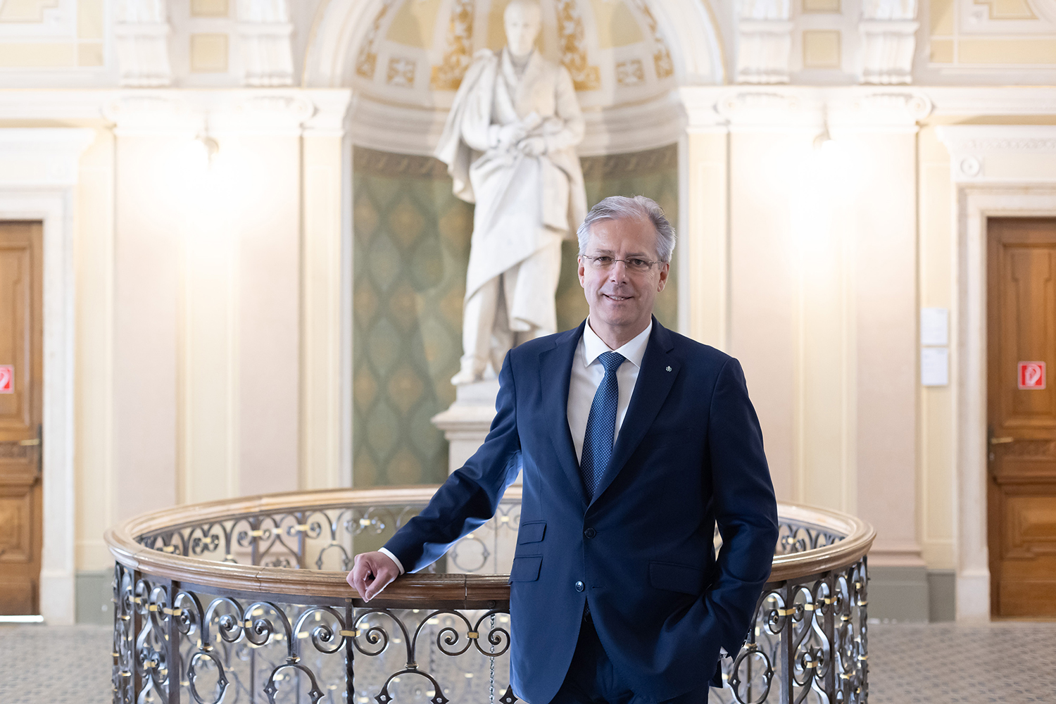 Ein Mann im Anzug steht an einem kunstvollen Geländer in einem eleganten Innenraum, mit einer großen weißen Statue und gewölbten architektonischen Details im Hintergrund.