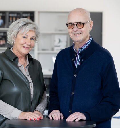 Eine ältere Frau und ein älterer Mann stehen in einem Haus zusammen und lächeln in die Kamera. Die Frau hat graues Haar und trägt ein graues Hemd über einer grauen Bluse; der Mann hat eine Brille und trägt einen blauen Pullover über einem Kragenhemd. Im Hintergrund sind Regale zu sehen.