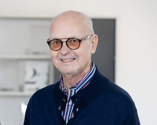 Ein älterer Mann mit Brille, einer Glatze und einem freundlichen Lächeln trägt einen dunklen Pullover über einem bunt gestreiften Hemd. Er steht drinnen in einem hellen, modernen Raum mit unscharfen Regalen im Hintergrund.