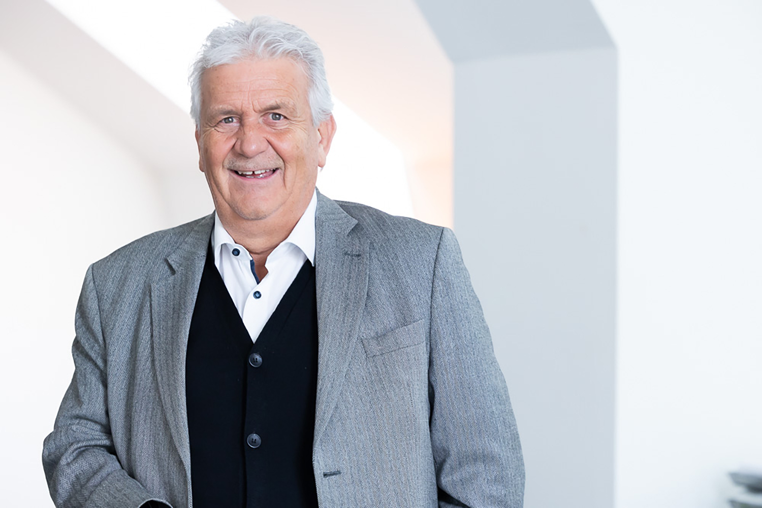 Ein älterer Mann mit weißem Haar, grauem Anzug, weißem Hemd und schwarzem Pullover steht lächelnd in einem hellen, modernen Innenraum mit weißen Wänden.