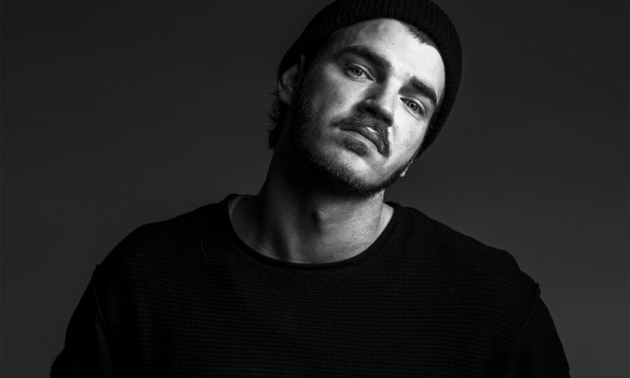 Ein Mann mit schwarzer Mütze und dunklem Pullover neigt den Kopf leicht und blickt mit neutralem Blick in die Kamera vor einem dunklen, schlichten Hintergrund. Das Bild ist in schwarz-weiß gehalten.