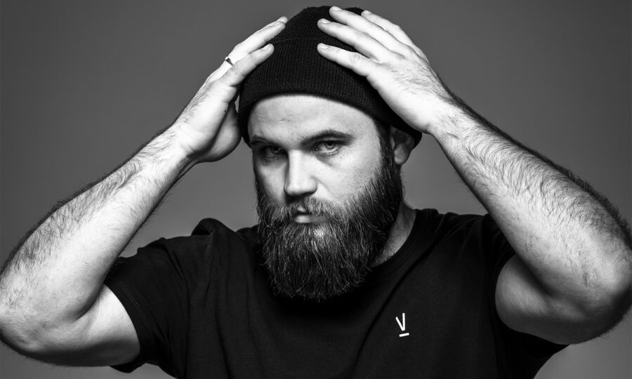Ein bärtiger Mann, der eine schwarze Mütze und ein schwarzes T-Shirt trägt, steht vor einem schlichten Hintergrund und blickt mit ernster Miene in die Kamera, während er beide Hände auf den Kopf legt.