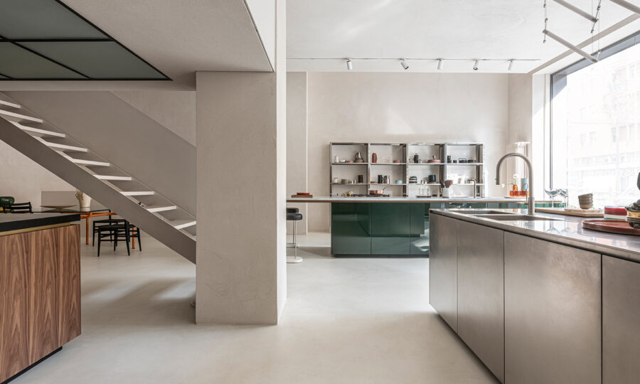 Moderne, minimalistische Kücheneinrichtung mit eleganten Schränken, Edelstahloberflächen, offenen Regalen mit Geschirr, einem großen Fenster für natürliches Licht und einer Treppe, die in ein höheres Stockwerk führt. Neutrale Töne und klare Linien dominieren den Raum.