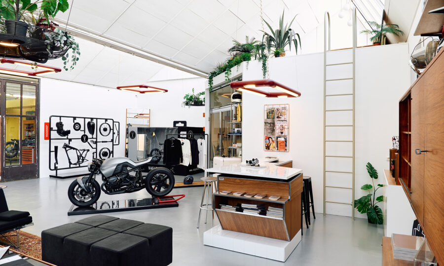 Ein moderner, offen gestalteter Arbeitsbereich mit einem eleganten schwarzen Motorrad, weißen Wänden, hängenden Pflanzen, einem zentralen Inseltisch, modernen Möbeln, Wandkunst und einer Leiter, die zu einem Loftbereich führt.