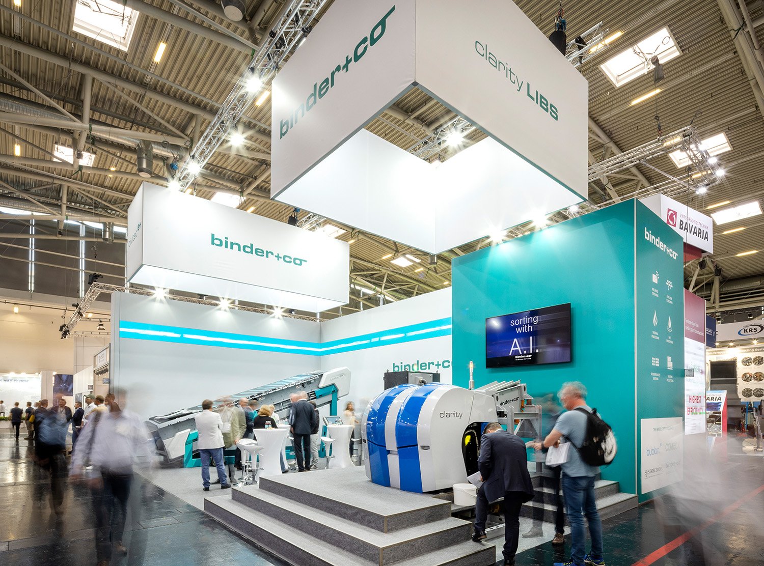 Ein moderner Messestand für binder+co auf einer Fachmesse, mit einer großen Maschine, Informationsdisplays, Sitzgelegenheiten und Besuchern, die in dem hell beleuchteten, industriell anmutenden Raum interagieren und umhergehen.