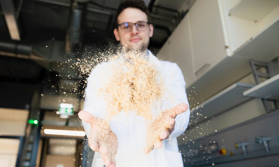 Eine Person in einem weißen Laborkittel sprüht Sägemehl oder feine Partikel in die Luft in einer Laborumgebung, in der im Hintergrund Regale und Industrierohre zu sehen sind.