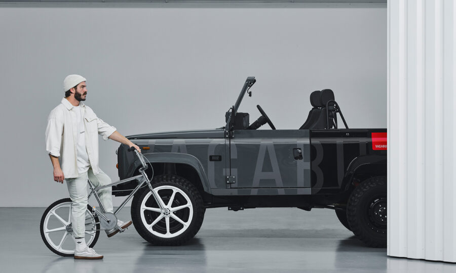 Ein weiß gekleideter Mann mit einer Mütze steht in einem Innenraum neben einem schwarzen Cabrio-Geländewagen und hält ein silbernes BMX-Fahrrad neben einer weißen Trennwand.