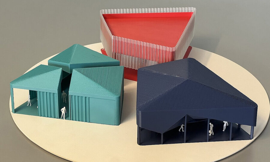 Auf einer runden Plattform sind vier architektonische Modelle zu sehen: drei blaugrüne und marineblaue Dreiecksstrukturen mit kleinen, weißen menschlichen Figuren im Inneren und ein durchscheinendes Dreieck mit roter Spitze im Hintergrund.