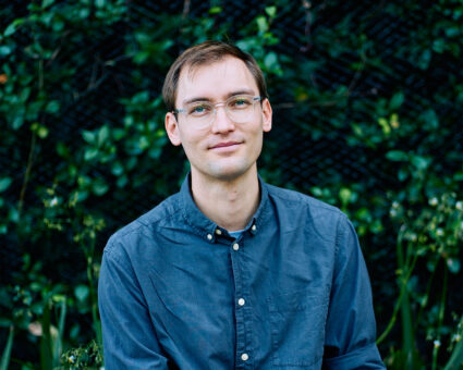 Ein junger Mann mit Brille und kurzen braunen Haaren, der ein blaues Button-up-Hemd trägt, sitzt im Freien vor einem grünen, begrünten Hintergrund.