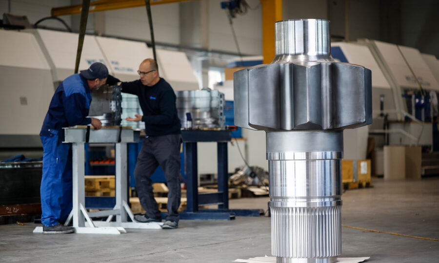 Ein großes, glänzendes Metallgetriebe steht auf dem Boden einer Fabrik. Im Hintergrund besprechen zwei Arbeiter in blauen Uniformen Papierkram in der Nähe von Industriemaschinen.