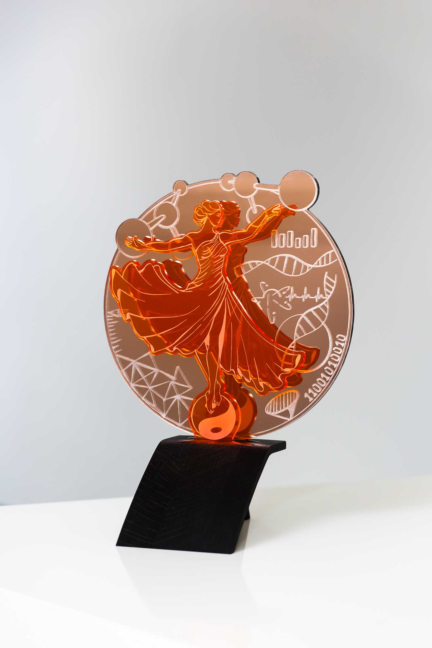 Ein orangefarbener Acryl-Award mit einer Frau in einem fließenden Kleid, die Kugeln hält, mit wissenschaftlichen Symbolen wie DNA, Diagrammen und Formeln im Hintergrund, ausgestellt auf einem schwarzen Ständer.
